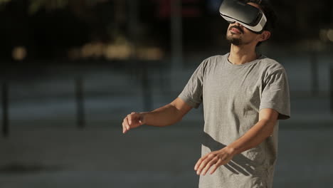 Young-Arabic-man-with-dark-curly-hair-and-beard-in-grey-T-shirt-being-in-park-in-virtual-reality-glasses